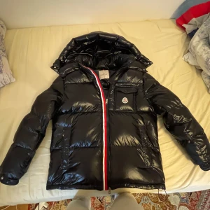 Svart dunjacka från Moncler - !ENDAST MÖTS UPP I MALMÖ GÅR EJ POSTA OSV TAR BARA KONTANTER!!Säljer en svart, glansig dunjacka från Moncler med huva och klassisk Moncler-logga på bröstfickan. Jackan har lång ärm, dragkedja med röd, vit och blå detalj samt flera fickor. Perfekt för kalla dagar och riktigt snygg streetstil.