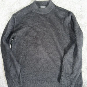 Svart stickad polotröja - Säljer en enkel och stilren svart stickad polotröja med lång ärm. Tröjan har ribbade muddar vid ärmslut och nederkant samt en hög krage. Perfekt för kyligare dagar.