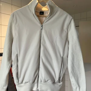 Vit bomberjacka från Porsche x BOSS - Säljer en stilren vit bomberjacka från Porsche BOSS med dragkedja och två fickor framtill. Jackan har ribbade muddar och ett stort Porsche BOSS-märke på ärmen. Perfekt för dig som gillar snygga detaljer.