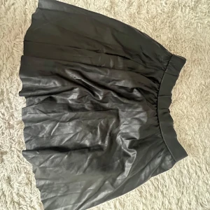 Svart kjol  - Snygg svart kort kjol i skinnimitation med lätt utställd form och bred midja. Perfekt för dig som vill ha en edgy och trendig look. Kjolen har en glansig finish och passar till många olika stilar.