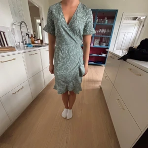 Grön prickig omlottklänning - Söt grön kortklänning med vita prickar och volangkant nertill. Omlottmodell med v-ringning och knyt i midjan. Kortärmad och lätt material, perfekt för varmare dagar.