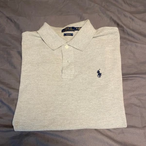 Ljusgrå pikétröja från Polo Ralph Lauren - Säljer en ljusgrå pikétröja från Polo Ralph Lauren, splitter ny i modellen Custom Fit. Tröjan har klassisk krage, knappar vid halsen och den ikoniska broderade loggan i marinblått på bröstet. Perfekt för en stilren och avslappnad look.