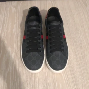 Svarta gucci ace skor - Säljer ett par snygga Gucci sneakers i mörkgrått med klassiskt monogrammönster och röd/blå rand på sidan. Baksidan har detaljer i rött och blått med Gucci-logga. Skorna har vit sula och svarta skosnören. Perfekt för dig som gillar lyxiga detaljer.