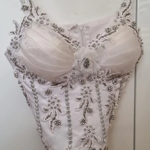 Vit dekorerad korsett - Superfin vit långklänning med glittriga detaljer, blommiga broderier och pärlor. Klänningen har smala axelband och en vacker blomapplikation framtill. Perfekt för speciella tillfällen när du vill sticka ut.