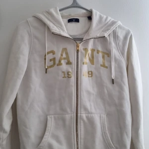 Vit hoodie med dragkedja från GANT - Säljer en vit hoodie från GANT med dragkedja och huva. Tröjan har guldiga bokstäver och siffror framtill samt två fickor. Perfekt för en avslappnad stil och enkel att matcha med det mesta. Oanvänd köpt i new york för 700