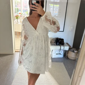 Vit spetsklänning med v-ringning - Säljer en romantisk vit kortklänning med v-ringning och långa, transparenta ärmar. Klänningen har vackra spetsdetaljer och öppen rygg med knytband. Perfekt för sommarens tillfällen!