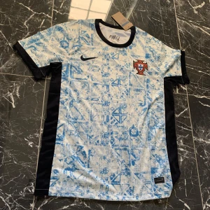 Portugal fotbollströja Nike blå/vit - Säljer en Portugal fotbollströja från Nike med unikt blått mönster på vit bas. Tröjan har svarta paneler längs sidorna och är kortärmad. Perfekt för dig som vill sticka ut på planen eller på läktaren.