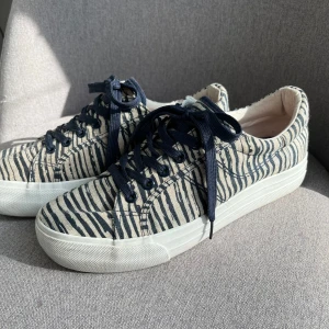 Randiga sneakers med marinblå snörning - Säljer ett par snygga sneakers med marinblå och vita ränder samt matchande marinblå snörning. Skorna har en vit platt sula och rund tå. Perfekta för dig som gillar en stilren men ändå unik look.