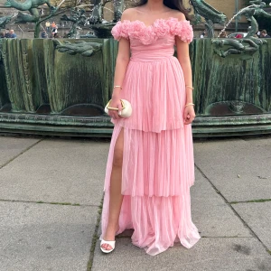 Rosa balklänning  - Romantisk ljusrosa långklänning med flera lager tyll, offshoulder-modell och stora volangdetaljer vid bysten. Klänningen har slits på ena sidan och markerad midja för en feminin siluett. Perfekt för bal eller fest. Hade denna underbart fina på min bal, och fick tusentals komplimanger!! Verkligen sjukt vacker klänning, men kommer inte ha någon nytta av den mer. Köpte denna för 2000 kr, och haft den endast en gång.