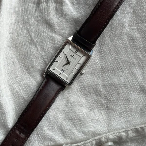 Limited edition klocka från Morris med brun läderrem - Stilren klocka från Morris med rektangulärt silverfärgat boett och vit urtavla. Klockan har en mörkbrun läderrem och minimalistiska detaljer. Perfekt accessoar för en klassisk look. Batteriet behövs bytas men annars i mycket bra skick.