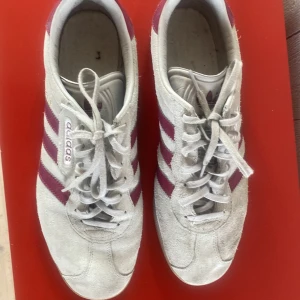 Adidas sneakers med vinröda/rosa ränder - Säljer ett par klassiska Adidas sneakers i vitt mocka med vinröda/rosa ränder och detaljer. Skorna har snörning, gummisula och den ikoniska Adidas-loggan på hälen. Perfekta för dig som gillar retrostil och streetwear. Den är tyvärr lite skadad på hälen. Har inga sulor heller så det får man köpa till. 