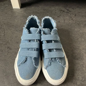 Blå jeanssneakers med fransade kanter - Säljer ett par blå sneakers i jeansmaterial från Even&Odd. Skorna har fransade kanter och tre breda kardborreband över foten. Vit, platt sula med mönstrad undersida. Perfekt för dig som gillar en unik och avslappnad stil. Använda 1-2 gånger så fortfarande i ett mycket bra skick. 