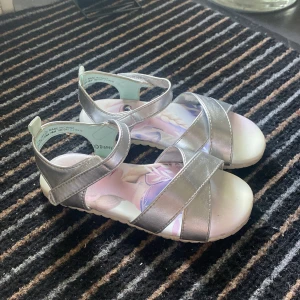 Silverfärgade sandaler med Frost-motiv - Säljer ett par glittriga silverfärgade sandaler med motiv av Elsa och Anna från Disney Frost på innersulan. Skorna har breda remmar och kardborreknäppning för enkel påtagning. Perfekta för sommaren och för alla små Frost-fans! Storleken är 26 och passar super bra till småbarn