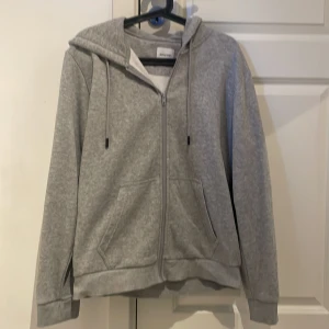 Grå hoodie med dragkedja från Jack & Jones - En stilren grå hoodie från Jack & Jones med dragkedja framtill och två fickor. Tröjan har huva med snörning och ribbade muddar vid ärmslut och nederkant. Perfekt för en avslappnad look.