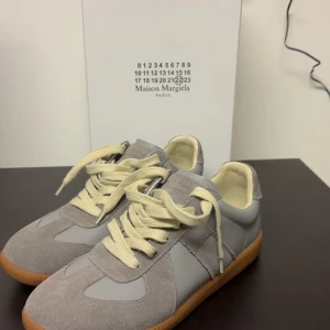 Grå sneakers från Maison Margiela - Säljer ett par stilrena grå sneakers från Maison Margiela med beige snörning och gummisula. Skorna har en mix av mocka och skinn, klassisk design och neutrala färger som passar till mycket.