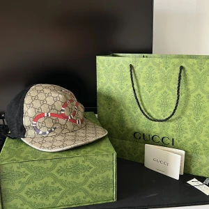 Gucci keps med orm-motiv - Snygg keps från Gucci med klassiskt GG-mönster i beige och brunt, dekorerad med en broderad röd och vit orm på framsidan. Baksidan är i svart mesh och justerbar rem med grönt och rött band. Perfekt accessoar för en trendig look.
