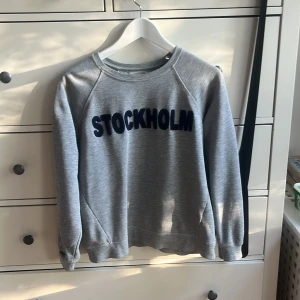 Grå sweatshirt med STOCKHOLM-tryck - Säljer en grå sweatshirt med mörkblå texten 'STOCKHOLM' på bröstet. Tröjan har rund halsringning och långa ärmar. Perfekt för en avslappnad stil.