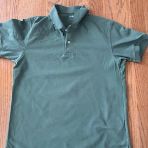 Grön pikétröja Uniqlo - Säljer en klassisk grön pikétröja från Uniqlo med krage och två knappar framtill. Tröjan är kortärmad och har en stilren, enkel design som passar perfekt till sommaren. Tillverkad i bomull för en bekväm känsla.