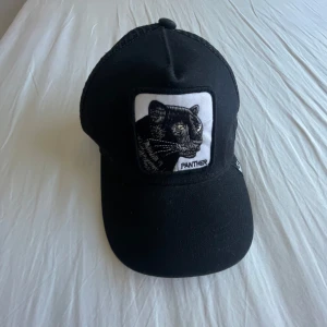 Svart keps med pantermärke - Svart keps med böjd skärm och ett stort tygmärke framtill med en broderad panter och texten 'Panther'. Kepsen har klassisk passform och justerbar baksida. Perfekt för dig som gillar djurmotiv och streetstyle.