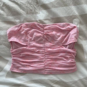 Rosa tubtopp med rynkade detaljer - Säljer en söt rosa tubtopp med rynkade sömmar framtill. Toppen är axelbandslös och har en mjuk, stretchig passform. Perfekt för varma dagar eller som ett lager under andra plagg.