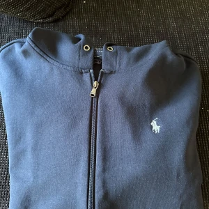 Mörkblå hoodie med dragkedja från Polo Ralph Lauren - Mörkblå hoodie med dragkedja från Polo Ralph Lauren. Klassisk design med huva, fickor och den ikoniska broderade loggan på bröstet. Perfekt för en avslappnad stil.