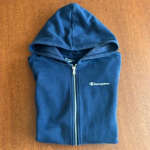 Mörkblå hoodie från Champion - Säljer en mörkblå hoodie från Champion med dragkedja framtill och huva. Tröjan har två fickor och ett vitt Champion-tryck på bröstet. Perfekt för en avslappnad stil. lite urtvättad.