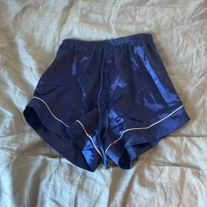 Blå glansiga pyjamas shorts med vit kant - Säljer ett par blå glansiga pyjamas shorts med vit kant och elastisk midja. De har en lös passform och är perfekta för sommarens varma dagar. Materialet känns svalt och lätt mot huden.