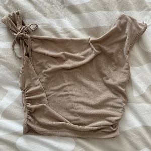 Beige oneshoulder topp med knyt - Trendig beige oneshoulder topp med knytning på axeln och rynkade detaljer längs sidan. Tillverkad i mjukt material som känns skönt mot huden. Perfekt för dig som vill sticka ut med en unik och stilren look.