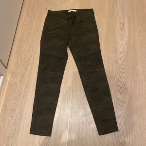 Mörkgröna camouflagebyxor med dragkedjedetaljer - Snygga mörkgröna byxor med camouflage-mönster och dragkedjor framtill. Byxorna har tight passform och ribbade detaljer vid knäna för en cool look. Perfekta för dig som vill sticka ut lite extra.