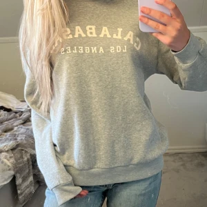 Grå sweatshirt från SHEIN  - Säljer en grå sweatshirt med vitt tryck där det står 'Calabasas Los Angeles' på bröstet. Tröjan har rund halsringning och långa ärmar med ribbade muddar. Perfekt för en avslappnad och stilren look.