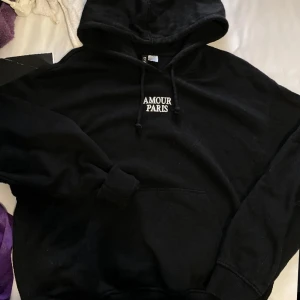 Svart hoodie med texten 'AMOUR PARIS' - Svart hoodie med luva och dragsko, samt vit broderad text 'AMOUR PARIS' på. Skön och fin.