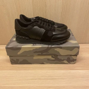 Valentino Rockrunner sneakers i svart med kamouflagemönster - Säljer ett par Valentino Rockrunner sneakers i svart med diskret kamouflagemönster. Skorna har svarta snören, platt sula och detaljerade nitar på hälen. Perfekta för dig som gillar exklusiva och stilrena sneakers. Pris ej skrivet i sten😊Kartong och tillbehör ingår
