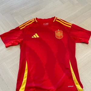 Spansk landslagströja från Adidas - Säljer en röd fotbollströja från Adidas med Spaniens landslagsemblem på bröstet. Tröjan har gula detaljer och klassiska Adidas-ränder på axlarna. Tillverkad i lätt material med AEROREADY-teknologi för bra ventilation. Perfekt för fotbollsträning eller match.