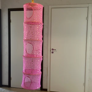 Rosa hängande förvaring med stjärnmönster - Säljer en rosa hängande förvaring med flera fack. är dekorerad med guldiga stjärnor och månar och har rundade öppningar på varje nivå. Perfekt för att organisera småsaker eller gossedjur på ett snyggt sätt.