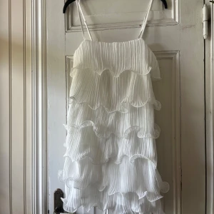 Vit volangklänning  - Säljer en vit volangklänning med tunna axelband och flera lager volanger i plisserat tyg. Klänningen har en luftig och romantisk känsla, perfekt för sommarens tillfällen. Supersöt och trendig modell!