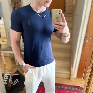 Mörkblå t-shirt från Polo Ralph Lauren - Säljer en mörkblå t-shirt från Polo Ralph Lauren i modellen Custom Slim Fit. T-shirten har korta ärmar, rund halsringning och en liten broderad logga på bröstet. Perfekt för en stilren och enkel look.