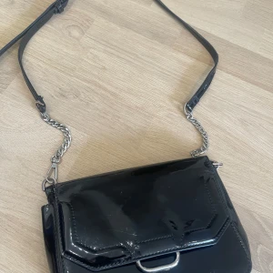 Svart axelväska i lack med silverkedja - Säljer en svart axelväska i glansigt lackmaterial med silverfärgad kedja och detaljer. Väskan har ett stilrent och modernt utseende med innerfack och dragkedja. Perfekt för att lyfta din outfit.