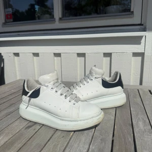Vita sneakers från Alexander McQueen - Säljer ett par klassiska vita sneakers från Alexander McQueen med svart detalj på hälen och tjock vit plattformssula. Skorna har rund tå, snörning och diskreta hål på sidorna för ventilation. Perfekta för en stilren look. Ingen box 