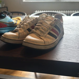 Vita Adidas sneakers med röd och svart rand - Säljer ett par klassiska vita Adidas sneakers med svarta och röda ränder på sidorna. Skorna har snörning och rund tå, perfekt för en sportig och stilren look. Adidas-logga på plösen och vit platt sula.