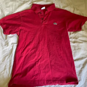 Röd piké t-shirt från Lacoste - Säljer en klassisk röd piké t-shirt från Lacoste med krage och knappar framtill. Ikoniska krokodilloggan broderad på bröstet. Perfekt för en stilren och sportig look.