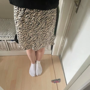 Beige och svart mönstrad kjol - Säljer en beige kjol med svart zebramönster. Kjolen har rak passform och är knälång, perfekt för dig som gillar att sticka ut lite extra. Materialet känns mjukt och följsamt mot huden. Supersnygg att styla med en enkel topp eller hoodie.