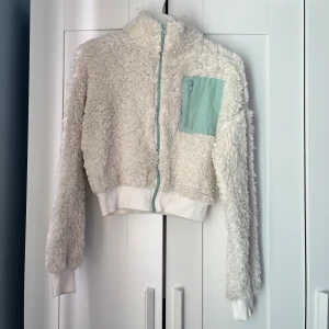 Vit teddyjacka med mintgrön ficka - Vit croppad teddyjacka med hög krage och mintgröna/blåa detaljer från Nelly, storlek XS, använd fåtal gånger. Bra höst/vår jacka!