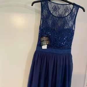 Marinblå paljettklänning med spets från New Look - Säljer en marinblå klänning från New Look med glittriga paljetter och spets upptill. Klänningen har rund halsringning, är ärmlös och har ett markerat midjeband. Kjolen är lätt utställd och slutar ovanför knäna. Klänningen är helt ny med prislappen på.