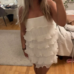 Vit volangklänning med plisserade lager - Säljer en söt axelbandslös vit klänning med flera plisserade volanglager. Klänningen är kort och har en luftig känsla, perfekt för sommarens tillfällen. Enkel att styla och ger en fräsch look. Aldeig använd men ingen prislapp på