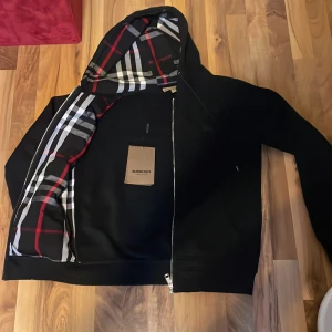Svart kofta/hoodie/jacka från Burberry - Svart hoodiejacka från Burberry med dragkedja och klassiskt rutigt foder i huvan. Jackan har fickor framtill och diskret broderad logga på bröstet. Perfekt för en stilren och avslappnad look.