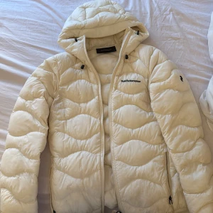 Vit dunjacka med huva från Peak Performance - Säljer en vit dunjacka från Peak Performance i storlek XS. Jackan har huva, dragkedja och diskret logga på bröstet. Perfekt för kyliga dagar och har en klassisk pufferstil med quiltat mönster.