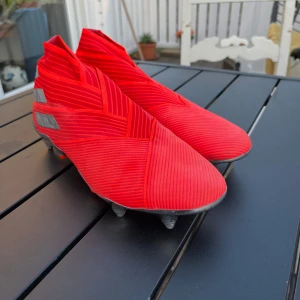 Röda Adidas Nemesis fotbollsskor med skruvdobb - Säljer ett par röda Adidas Nemesis med skruvdobb och med silvriga detaljer och mönstrad ovandel. Skorna har en strumpliknande passform utan snörning och en glänsande sula med fasta dobbar. Perfekta för dig som vill sticka ut på planen! De är i princip oanvända och sitter bra på foten. Är öppen för förhandlingar 