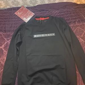 Svart sweatshirt från Hugo Boss - Svart långärmad sweatshirt från Hugo Boss med klassisk rund hals och tydlig logotyp framtill. Tröjan har en stilren design och är tillverkad i mjuk bomullsblandning som känns skön mot huden. Perfekt för dig som gillar en clean och exklusiv look.