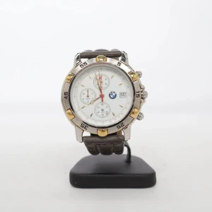 BMW herrklocka med brun läderrem - Stilren BMW-klocka med vit urtavla, silverfärgat boett och guldiga detaljer. Klockan har tre små urtavlor, datumvisning och en brun vadderad läderrem. Perfekt accessoar för dig som gillar klassisk design och bilar.        Behöver bara ett nytt batteri.