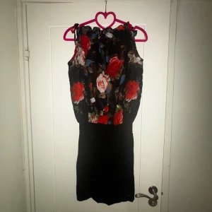 Svart kortklänning med blommigt mönster - Snygg svart kortklänning med blommigt mönster i rött, rosa och vitt upptill. Klänningen är ärmlös och har en loose passform upptill med knyt vid axlarna. Perfekt för dig som gillar färgstarka detaljer och vill sticka ut lite extra.